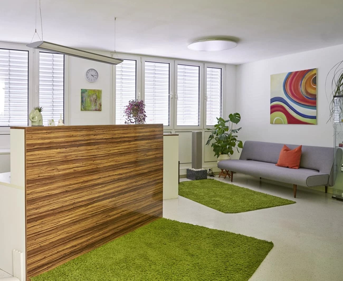 Moderner Empfangsbereich einer Praxis mit hölzernem Tresen, grünem Teppich, grauem Sofa und hellen Fensterfronten.